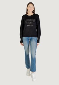 Schwarzer Pullover mit "GUESS"-Logo aus Strass, kombiniert mit hellblauen Schlagjeans und weißen Sneakers, vor einem neutralen Hintergrund.