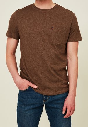 Uomo che indossa una t-shirt marrone a maniche corte con taschino sul petto e jeans blu, mano in tasca, su sfondo chiaro uniforme.