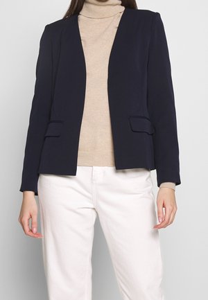 Femme portant un blazer bleu marine par-dessus un pull à col roulé beige et un pantalon blanc, debout devant un fond uni.