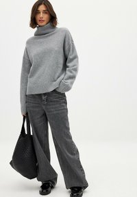 Gray knit turtleneck sweater paired with loose gray denim jeans. Model holds a black woven tote bag. Glossy black shoes complete the look.