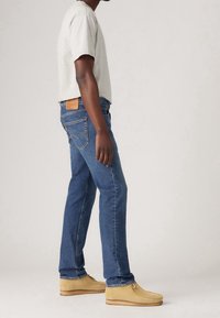 Blue denim jeans with a straight-cut design, tan leather back patch, and beige moccasin-style shoes; grey crew-neck t-shirt worn.