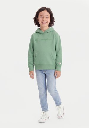Smilende barn med kort mørkt hår iført en grøn Champion hættetrøje, lyseblå jeans og hvide høje sneakers, stående på en hvid baggrund.