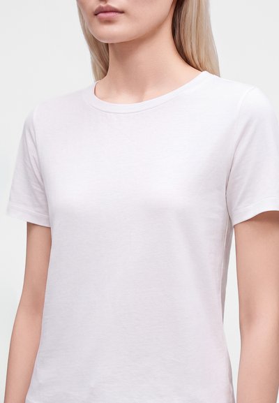 Jeune personne à la peau claire et aux longs cheveux blonds portant un t-shirt blanc uni à manches courtes et col rond sur un fond blanc.