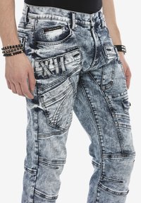 Ausgebleichte blaue Denim-Jeans mit strukturiertem Finish; verfügen über mehrere Taschen und dekorative Nähte mit "XII"-Details auf der Vorderseite.