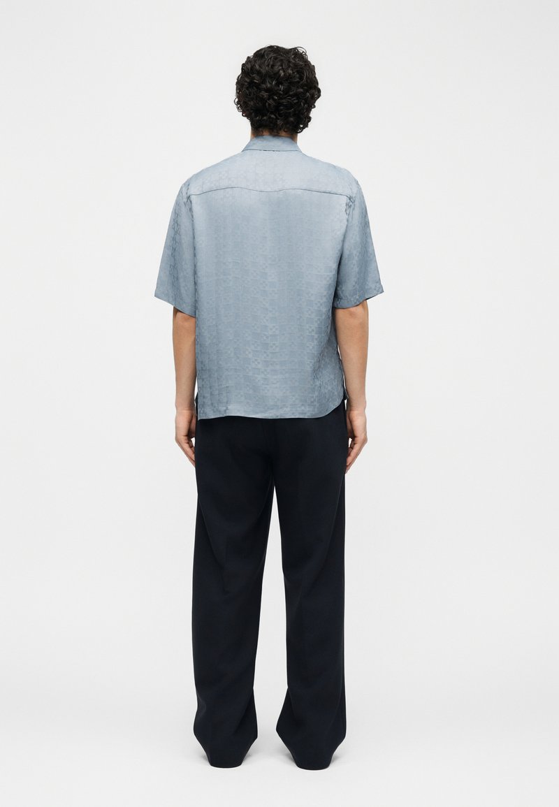 Man with curly hair stands facing away wearing a short-sleeve light blue shirt and loose black pants against a plain background.