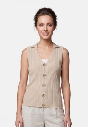 Giovane donna con capelli castani, indossa un top beige senza maniche a coste con bottoni dorati unici e pantaloni beige chiaro, sorridente su sfondo uniforme.