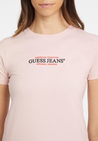 Camiseta de algodón de color rosa claro con cuello redondo, que muestra un texto del logo "GUESS JEANS" en negro y rojo y "AMERICAN TRADITION" encima.