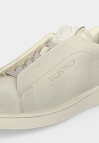 Gros plan d’une basket Valentino en cuir blanc cassé avec logo embossé, surface texturée et lacets beige clair.