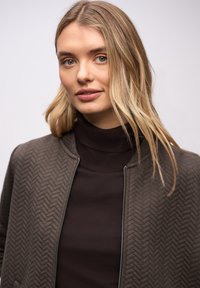 Gewatteerd bomberjack in donkerolijfgroen met een gestructureerd visgraatpatroon, gecombineerd met een bruine coltrui. Ritssluiting aan de voorkant en ronde kraag.