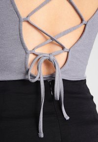 Šedý žebrovaný top s nízkým výstřihem na zádech a křížovými ramínky, se zavazovacím detailem. Černá látka viditelná na spodní části se zipem.