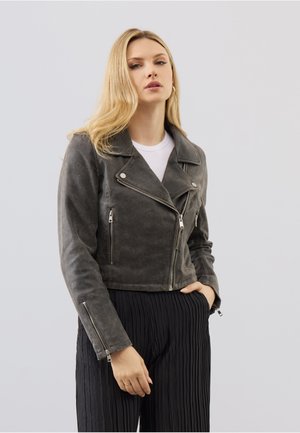 Frau mit langen blonden Haaren, die eine graue Wildleder-Bikerjacke über einem weißen Top und schwarzen plissierten Hosen trägt und mit einer Hand in der Tasche steht.