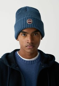 Modrá pletená čiapka z mäkkej priadze s okrúhlym štítkom s logom "COLMAR". Nosí sa s modrým svetrom a tmavou bundou. Textúrovaný rebrovaný vzor.