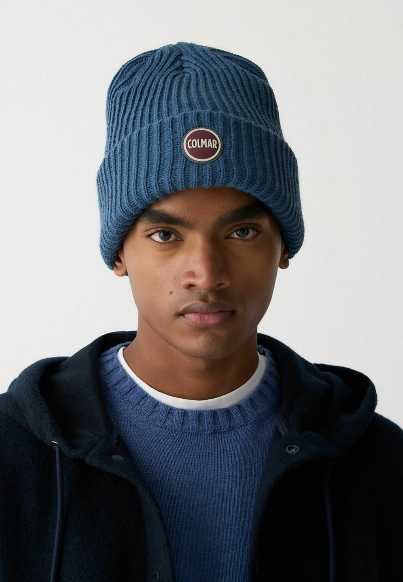 Modrá pletená čiapka z mäkkej priadze s okrúhlym štítkom s logom "COLMAR". Nosí sa s modrým svetrom a tmavou bundou. Textúrovaný rebrovaný vzor.