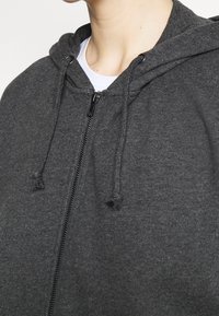 Grijze ritshoodie van zachte stof met een capuchon, trekkoorden en een soepele rits. Heeft een ontspannen pasvorm en een minimale textuur.