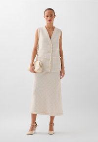 Crème tweed vest met franje randen, knoopsluiting en twee zakken, gecombineerd met een bijpassende midi-rok. Clutch in de hand.