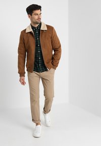 Brun nubuck bomberjakke med en lysebeige faux shearling krave, stylet over en grønternet skjorte, parret med beige bukser og hvide sneakers.