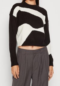 Svart och vit kortad tröja i mjukt stickat material, med abstrakta vågmönster. Långa ärmar och rund halsringning. Kombinerad med gråa byxor.