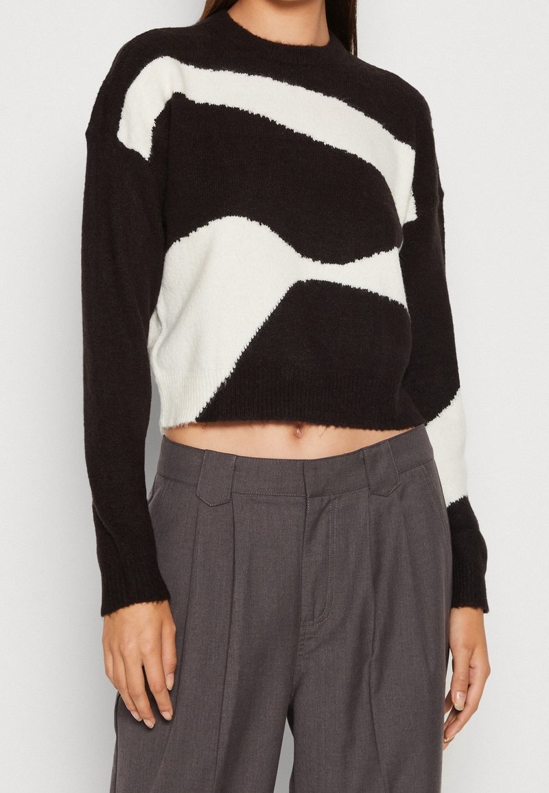 Svart och vit kortad tröja i mjukt stickat material, med abstrakta vågmönster. Långa ärmar och rund halsringning. Kombinerad med gråa byxor.