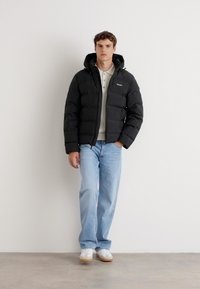 Zwarte pufferjack met een capuchon, voorzien van vierkante secties en een klein logo. Gecombineerd met lichtblauwe jeans en witte sneakers.