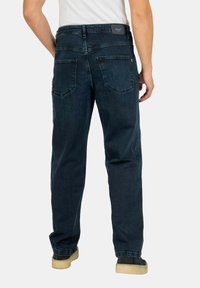 Vaqueros de mezclilla azul oscuro con un corte recto, que cuentan con dos bolsillos traseros y una etiqueta de cuero en la cinturilla. Llevados con zapatos beige.