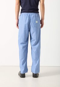 Asmuo, dėvintis mėlynas ir baltas vertikaliai dryžuotas kelnes su užpakaline kišene ir juodus batus, stovintis prieš paprastą baltą foną.