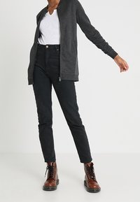 Persona che indossa una maglietta bianca a V, una giacca zip-up grigio scuro, jeans aderenti neri a vita alta e stivali marroni con lacci, in piedi su uno sfondo bianco.