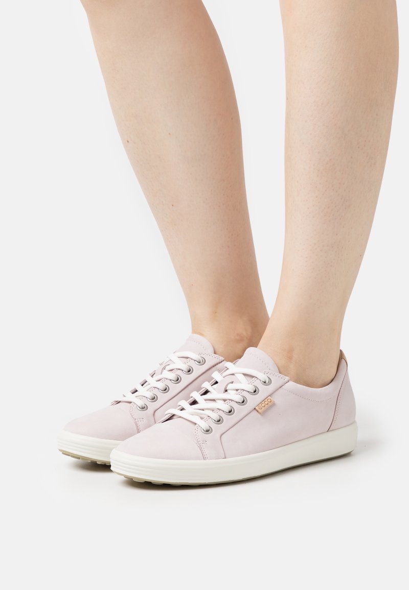 Personne portant des baskets à lacets rose clair avec des semelles blanches, debout sur un fond blanc uni.