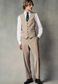 Costume beige clair avec un gilet ajusté à cinq boutons, un pantalon à devant plat et une chemise blanche. Accompagné d'une cravate verte foncée et de chaussures noires.