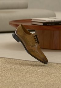 Καφέ δερμάτινο παπούτσι φόρμας με υφή κροκοδείλου να αιωρείται πάνω από μπεζ χαλί σε μοντέρνο καθιστικό με ξύλινο τραπέζι και βιβλία.