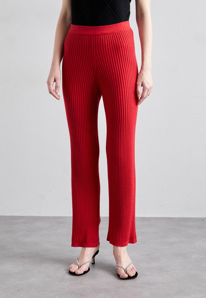 Rote gerippte Strickhosen mit hohem elastischem Bund, geradem Beinverlauf und fransigem Saum an den Knöcheln, kombiniert mit schwarzen Flip-Flops.