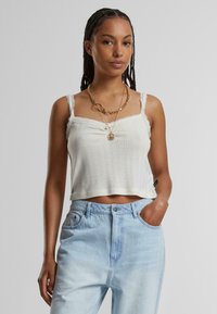 Kort, off-white ribbad linne med spetsdetaljer och smala axelband; kombinerat med ljusblå jeans med hög midja och lager av gyllene halsband.