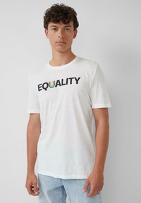 Junger Mann mit lockigem Haar, der ein weißes T-Shirt mit dem Aufdruck "EQUALITY" und einem regenbogenfarbenen U sowie hellblauen Jeans trägt, vor einem schlichten Hintergrund.