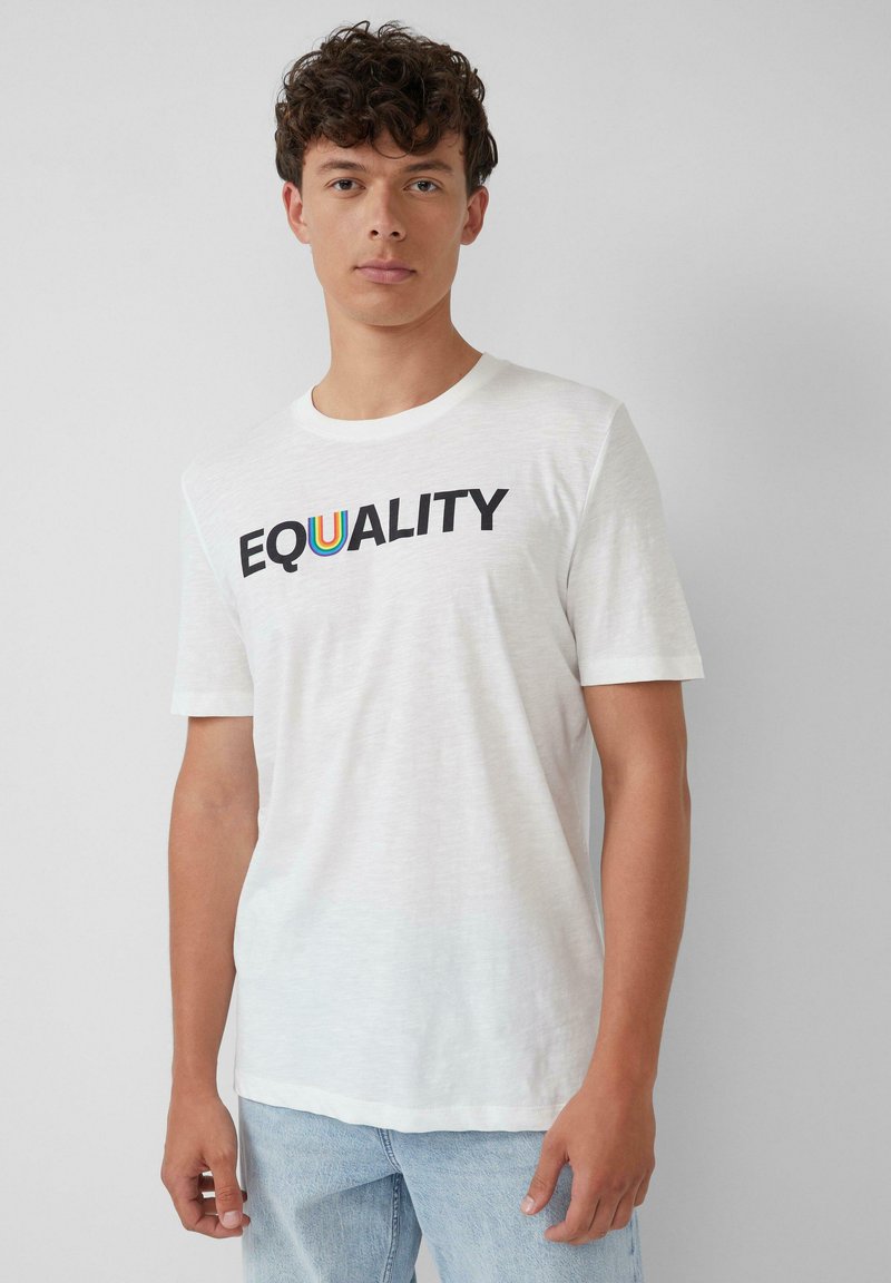 Junger Mann mit lockigem Haar, der ein weißes T-Shirt mit dem Aufdruck "EQUALITY" und einem regenbogenfarbenen U sowie hellblauen Jeans trägt, vor einem schlichten Hintergrund.