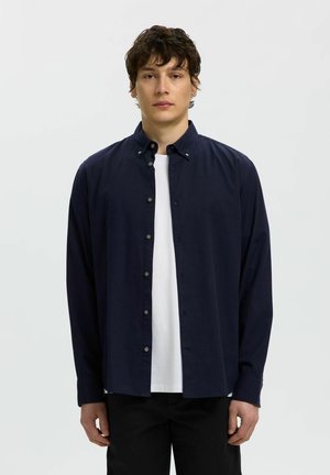 Junger Mann mit kurzen lockigen Haaren, der ein marineblaues Hemd über einem weißen T-Shirt trägt, steht vor einem schlichten hellen Hintergrund.