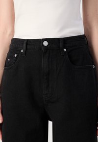 Mustat denim-farkut, joissa on korkea vyötärö, etutaskut ja hopeiset yksityiskohdat. Sileä rakenne ja perinteinen viiden taskun muotoilu.