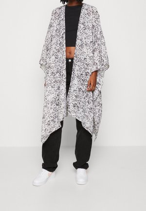 Personne portant un crop top noir, un pantalon noir ample, des baskets blanches et un kimono long blanc à pois gris ouvert sur le devant.
