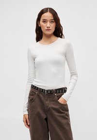 Ilgas baltas megztinis su dryžuota faktūra, derinamas su ruda aukšto liemens kelnėmis ir juodu diržu su sidabrinėmis ovaliomis skylelėmis.