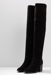 Botas de ante negras hasta la rodilla con tacón cuadrado, puntera puntiaguda y diseño sin costuras. Presenta una textura suave y un estilo minimalista.