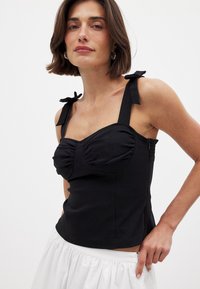 Top aderente nero realizzato in tessuto liscio con parte anteriore arricciata, spalline larghe e dettagli con lacci regolabili. Presenta un busto strutturato con arricciature laterali.