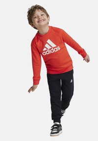 Sudadera roja con el logo blanco de Adidas, pantalones de chándal negros con detalles blancos y zapatillas negras con rayas blancas y suelas de goma.