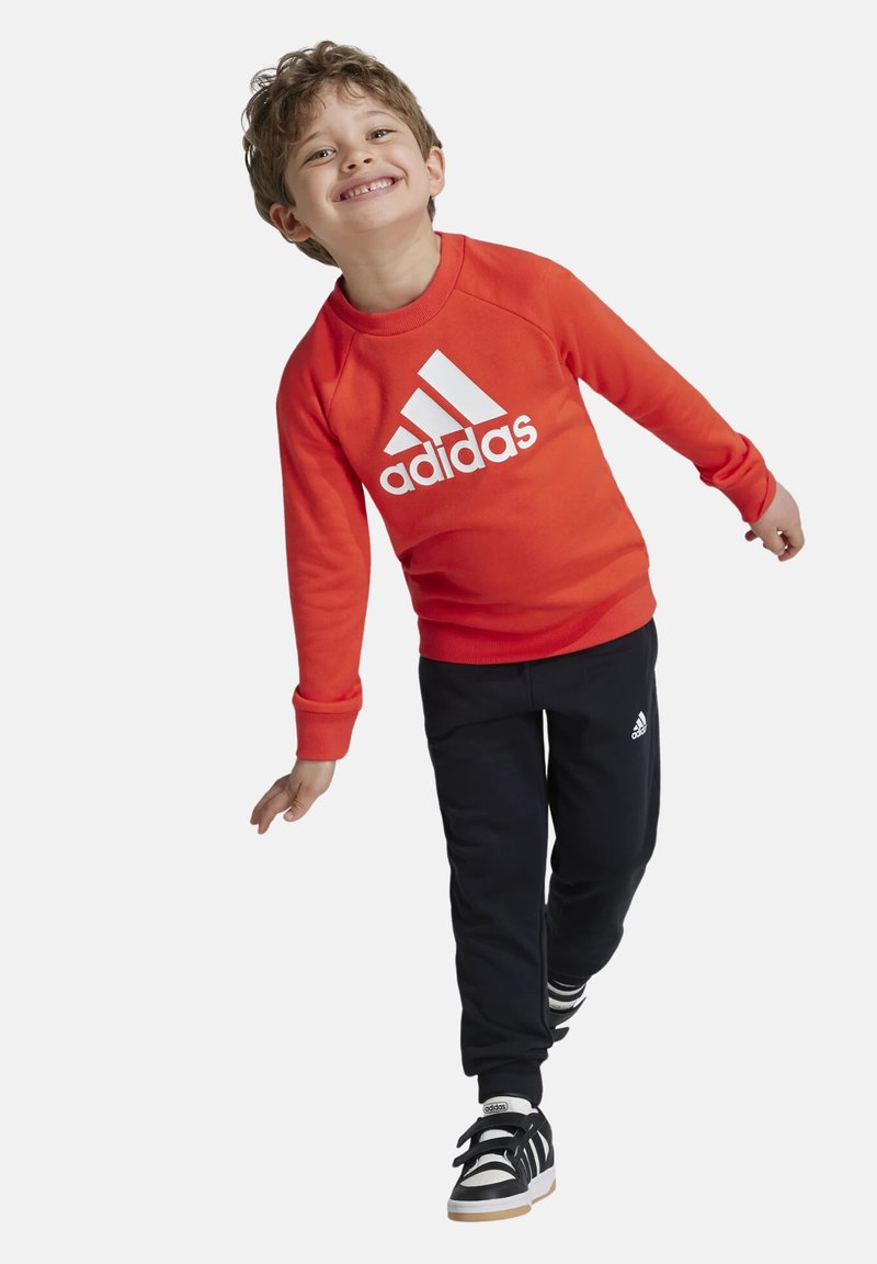 Sudadera roja con el logo blanco de Adidas, pantalones de chándal negros con detalles blancos y zapatillas negras con rayas blancas y suelas de goma.