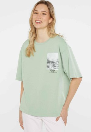 Young woman smiling, wearing light green oversized t-shirt with cloud print and text on front pocket area, paired with white pants.