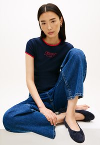 Μπλε ναυτικό κοντομάνικο μπλουζάκι με κόκκινες λεπτομέρειες και κεντημένο λογότυπο, συνδυασμένο με ανοιχτό μπλε παντελόνι με φαρδιά μπατζάκια και σκούρα μπλε επίπεδα παπούτσια.