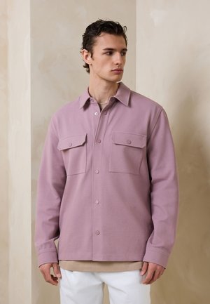 Joven con chaqueta de color púrpura claro abotonada con bolsillos en el pecho, camisa beige debajo y pantalones blancos, de pie frente a una pared beige.
