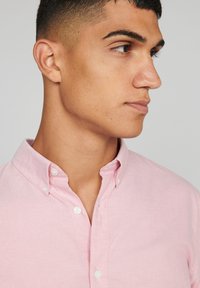 Lichtroze overhemd met knopen en een klassieke kraag, witte knopen en een gladde stofstructuur. Het ontwerp heeft een slim fit en minimale details.