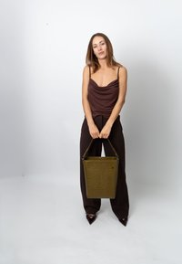 Olive green crocodile-patterned handbag with short strap. Model wears a brown cowl-neck top and dark wide-leg pants, standing in front of a plain background.