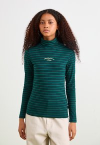 Grøn- og marineblå stribet turtleneck skjorte med lange ærmer, der har et lille gult logo på brystet. Blødt, strækbart stof.