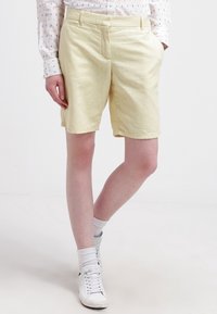 Shorts jaune clair jusqu'aux genoux en tissu doux avec un devant plat et des poches latérales, assortis à des baskets blanches et des chaussettes montantes.