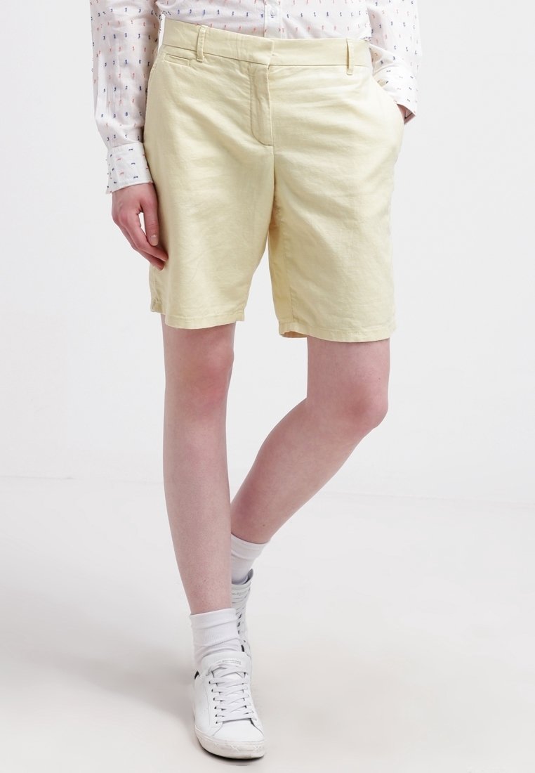 Shorts jaune clair jusqu'aux genoux en tissu doux avec un devant plat et des poches latérales, assortis à des baskets blanches et des chaussettes montantes.