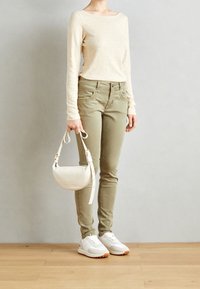 Lichtbeige top met lange mouwen, olijfgroene slim-fit broek, witte sneakers en een kleine witte handtas met een gebogen vorm.